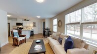 Located in Carrier Woods in Scarborough, Maine, this single-floor residence offers a thoughtfully designed open-concept layout that seamlessly connects the main living areas. The living room is bright and welcoming, anchored by large windows that flood the space with natural light and provide views of the surrounding neighborhood. Warm neutral tones on the walls complement the light hardwood-style flooring, creating a cohesive and inviting ambiance throughout.

The kitchen is clean and functional, featuring white cabinetry, light stone countertops, stainless steel appliances, and a peninsula with bar seating — ideal for casual dining or entertaining. Adjacent to the kitchen, a compact dining area with a bistro-style table and stools offers an additional space for meals.

The open flow between the kitchen, dining nook, and living room maximizes the sense of space, making the layout feel airy and versatile. Tasteful décor details, including woven textures, decorative pillows, and framed wall art, add character and warmth. The single-floor design ensures easy accessibility and convenient living, making this Carrier Woods residence a comfortable and practical home in the heart of Scarborough, Maine.