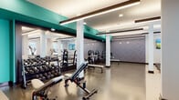 This expansive fitness center is thoughtfully designed to support a full range of workout routines across multiple distinct zones. The space features a clean, contemporary aesthetic defined by bold teal accent beams and columns contrasted against crisp white walls and ceilings. Durable epoxy and rubber flooring runs throughout, providing a practical and polished finish suited to high-traffic athletic use.

The free weights area is well-stocked with a comprehensive dumbbell rack and multiple adjustable benches, accompanied by a wall of mirrors that enhance the sense of space and allow for proper form monitoring. Adjacent to this zone, a dedicated cardio section houses a row of treadmills, ellipticals, and a stationary bike, positioned near windows that bring in natural light. Mounted flat-screen televisions provide entertainment throughout the facility.

A standout functional training room features an impressive array of equipment including battle ropes, resistance bands, medicine balls, kettlebells, and a multi-use cable machine. This room is accessed through an industrial-style roll-up door, adding a distinctive design element. A separate studio area equipped with stability balls and a wall-mounted display offers space for guided or independent training sessions. Recessed and linear LED lighting ensures the entire facility is bright and well-illuminated.