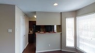 This single-story space is designed with an open-concept layout that seamlessly connects the kitchen and living areas. The kitchen is equipped with dark wood cabinetry and black appliances, including a refrigerator and range, creating a cohesive and modern aesthetic. A half-wall breakfast bar extends from the kitchen, providing a casual dining or serving surface that also defines the boundary between the two spaces without sacrificing openness.

The flooring throughout is dark hardwood, lending warmth and continuity to the overall design. Neutral beige walls complement the darker tones of the cabinetry and floors, creating a balanced color palette. A textured ceiling adds subtle visual interest overhead, while recessed lighting in the kitchen area provides focused illumination.

Natural light enters the living area through a window fitted with horizontal blinds, brightening the space during the day. The layout is practical and efficient, with a door visible near the kitchen that likely leads to additional storage or an entry point. Overall, the space presents a clean, functional design suited for everyday living.