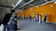 This contemporary fitness center is designed to deliver a motivating and fully equipped workout experience. The space is defined by its industrial-style aesthetic, featuring exposed concrete ceilings with visible ductwork and silver ventilation pipes running overhead, lending the room an open, loft-like atmosphere. Pendant lighting fixtures hang from the ceiling, providing focused illumination throughout the floor.

Bold orange accent walls energize the space and create a dynamic visual contrast against the neutral, textured carpet flooring. The layout is thoughtfully arranged to accommodate a comprehensive selection of strength training machines, including cable systems, seated press units, leg curl and extension machines, and a free weight rack with benches — offering a full range of resistance training options.

A wall-mounted flat-screen television is positioned for visibility across the workout area, adding an entertainment element for users during their sessions. The spacious floor plan ensures ample room between equipment, promoting both safety and comfort during use. The overall ambiance is modern and functional, combining an industrial design sensibility with the practical demands of a well-appointed fitness facility.