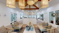 Located at 122 Pinewood Crescent in Yorktown, Virginia, this single-story facility offers a thoughtfully designed layout spanning a generous footprint across five distinct areas. The entrance greets visitors with a dramatic, soaring lobby anchored by exposed wood beam trusses and an eye-catching cluster of oversized drum pendant lights. Shiplap-clad white walls lend a coastal modern aesthetic, while patterned parquet-style hardwood flooring adds warmth and visual interest throughout. A central reception desk with a slatted vertical screen backdrop serves as the focal point of the welcoming area, flanked by comfortable seating arrangements ideal for gathering. Adjacent to the lobby, a spacious lounge area features built-in open shelving, a wall-mounted display, and large windows that invite abundant natural light and outdoor views. A dedicated meeting or breakout room offers a round wood table and floor-to-ceiling windows on two walls, creating a bright, productive environment with scenic views of the surrounding greenery. The fitness center is a standout space, finished with dark charcoal walls, a black ceiling, and polished concrete flooring. It is comprehensively equipped with cable machines, weight benches, dumbbells, kettlebells, and mirrored storage walls, providing a professional-grade workout environment within the facility.