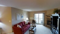 This single-floor residence offers a comfortable and inviting living environment designed with everyday functionality in mind. The living room serves as the centerpiece of the space, featuring plush wall-to-wall carpeting in a warm neutral tone that complements the soft, beige-toned walls throughout. A bold red sofa anchors the seating area, paired with a rattan-style armchair and ottoman, creating a cozy and relaxed atmosphere ideal for both lounging and entertaining. A wicker coffee table adds a natural, textured element to the room. Large sliding glass doors flood the living area with abundant natural light and provide direct access to a private balcony, offering views of the surrounding exterior. Sheer drapes frame the doors, softening the light while maintaining an open, airy feel. The room is finished with a textured ceiling and features tasteful wall art that adds a subtle decorative touch. Track lighting overhead provides additional illumination. A fireplace is also visible along one wall, contributing warmth and character to the overall ambiance. The layout is efficient and well-organized, making thoughtful use of the available space to create a residence that feels both functional and comfortably livable.