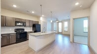 This 1-bedroom, 1-bathroom unit offers a thoughtfully designed single-floor layout that makes efficient use of its space. The open-concept kitchen and living area serves as the heart of the unit, where rich dark-toned shaker cabinetry, granite countertops with a subway tile backsplash, and black appliances — including a range, microwave, and refrigerator — define the kitchen. A central island with a built-in sink and pendant lighting above adds both functionality and style, flowing seamlessly into the living area. Light wood-style flooring runs throughout the main living spaces, complementing the neutral, warm wall tones. The living room features a ceiling fan and access to an outdoor balcony through a glass door flanked by tall windows, inviting generous natural light and outdoor views. The bedroom is a comfortable retreat with plush carpet, a ceiling fan, and a large window overlooking greenery. A dedicated closet and a hallway provide additional storage and connectivity within the unit. The full bathroom is clean and practical, featuring a soaking tub with a tiled shower surround, coordinating cabinetry, and wood-style flooring that ties into the overall aesthetic of the space.