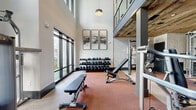 Located in Sugar Hill, GA, this two-story fitness center offers a comprehensive workout environment spread across a generously sized main floor and an upper-level mezzanine. The space blends modern design with rustic character, featuring reclaimed wood ceiling panels and accent walls alongside sleek concrete-style flooring and floor-to-ceiling windows that flood the interior with natural light and outdoor views.

The main floor is divided into dedicated workout zones. The cardio area is lined with a row of treadmills, stair climbers, and elliptical machines, complemented by large mirrors and linear LED lighting overhead. Adjacent to this is a strength training zone with rubber-floored areas housing multi-station cable machines, adjustable benches, and a full rack of dumbbells. Stone-textured accent walls add visual depth, while framed artwork contributes a polished, modern aesthetic.

Soaring double-height ceilings in the free weight area create an open, airy atmosphere, with pendant lighting and expansive windows enhancing the sense of volume. A staircase with metal railings leads to the upper mezzanine level, which provides additional functional space overlooking the main floor. Glass-paneled double doors connect distinct zones, maintaining an open flow throughout. The overall design balances industrial warmth with contemporary fitness facility standards.
