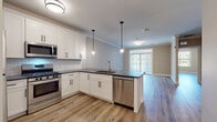 This single-floor space showcases a thoughtfully designed open-concept layout that seamlessly connects the kitchen, dining, and living areas. The kitchen is a standout feature, outfitted with crisp white shaker cabinets accented by matte black hardware, dark granite countertops, and a classic white subway tile backsplash. Stainless steel appliances — including a gas range, over-the-range microwave, dishwasher, and refrigerator — complete the well-equipped culinary space. Pendant lighting over the peninsula adds a modern touch, while a large window and glass French doors in the living area invite generous natural light and offer views of the surrounding greenery. Light wood-look flooring extends throughout the main living spaces, complemented by neutral warm-toned walls and crown molding that lend an air of refinement. The bedroom features its own large window with outdoor views and continues the light flooring aesthetic for a bright, airy feel. The bathroom presents a luxurious finish with large-format marble-style porcelain tile flooring and wall surround, a gray vanity with a white countertop, and a combined tub and shower. Overall, the space offers a clean, contemporary aesthetic with quality finishes throughout.