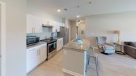 This single-floor unit presents a well-designed, open-concept layout with light wood-style flooring and neutral-toned walls that create a clean, cohesive aesthetic throughout. The kitchen serves as a central hub, showcasing white cabinetry, granite countertops, and stainless steel appliances including a gas range and full-size refrigerator. A curved kitchen island with an integrated sink and pendant lighting above provides both additional workspace and casual seating with metal bar stools. The open layout connects the kitchen seamlessly to the living area, which features a large sectional sofa, a round coffee table, and a wall-mounted television. Natural light enters through a front-facing window offering outdoor views. A secondary seating area includes a tufted wingback chair, a dark sofa, and a decorative table lamp with a patterned shade. The bedroom is furnished with a bed and a modern dark-toned dresser. Two bathrooms mirror each other in design, each featuring granite-topped white vanities, large mirrors with overhead lighting, tile flooring, and shower-tub combinations. The space maintains a consistent modern and functional character throughout.