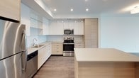 This well-designed one-bedroom, one-bathroom unit offers a clean, modern aesthetic across a single floor. The open-concept kitchen serves as a central highlight, featuring a large center island with a neutral quartz-style countertop, a mix of white gloss and light wood cabinetry, a soft blue glass tile backsplash, stainless steel appliances including a refrigerator, range, microwave, and dishwasher, and recessed lighting overhead. Dark hardwood-style flooring grounds the kitchen and flows into the adjoining living area.

The living space is bright and airy, anchored by a generous floor-to-ceiling window configuration that frames views of the surrounding urban streetscape and fills the room with natural light. The walls are crisp white throughout, contributing to a clean, contemporary feel.

The bedroom is carpeted for comfort and features its own large window with outdoor views, a walk-in closet with built-in shelving, and direct access to the rest of the unit. The bathroom is efficiently appointed with a single-sink vanity with light wood cabinetry, a combined tub and shower with a sliding glass door, and hardwood-style flooring that coordinates with the rest of the unit.