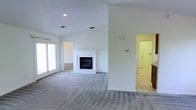This two-story home offers a well-organized layout with distinct living areas connected by thoughtful architectural design. The main living area is generously proportioned, featuring soft wall-to-wall carpeting and soaring ceilings that enhance the sense of openness and volume. A white-mantled fireplace serves as the room's focal point, adding a classic, welcoming element to the space. Large sliding glass doors along one wall allow abundant natural light to pour in, brightening the neutral white walls and contributing to the home's airy atmosphere.

A structural column divides the living area from an adjacent space, which transitions to light-colored tile flooring — likely a kitchen or utility area — visible through a connecting doorway. This tiled zone offers a practical contrast to the carpeted living space. Additional doorways suggest further rooms branching off the main area, providing a functional flow throughout the floor plan.

Recessed lighting in the ceiling supplements the natural light, ensuring the space remains well-lit at all times. The clean, neutral color palette of whites and grays throughout creates a versatile backdrop. The staircase visible near the tiled area provides access to the second floor, completing this multi-level home's layout.