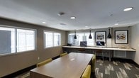 This single-floor shared workspace is designed to support productivity in a clean, contemporary setting. The room is anchored by a large rectangular conference or collaboration table surrounded by cushioned chairs upholstered in a muted yellow-green fabric, providing comfortable seating for group work or meetings. Overhead, sleek black pendant lights hang above the table, complementing the recessed ceiling lighting that evenly illuminates the space.

Along the perimeter wall, a continuous built-in desk runs beneath a series of windows that allow generous natural light to fill the room. This workstation area is equipped with desktop computers, offering dedicated spaces for individual tasks. The windows provide views of the surrounding building exterior, contributing to an open, connected atmosphere.

The flooring features a patterned carpet with geometric detailing, adding visual interest and texture underfoot. Walls are finished in a neutral gray tone, lending a professional yet approachable feel. A framed piece of wall art adds a subtle decorative touch. The overall layout balances communal collaboration space with individual workstations, making this a versatile and well-appointed shared amenity area.