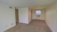 This single-floor space features an open layout connecting the main living area to a dining space. The floors throughout are covered in neutral beige carpet, contributing to a warm, understated aesthetic. The living area is generously proportioned, offering an open feel with white walls and a textured popcorn ceiling overhead.

A central dividing wall with bifold louvered closet doors provides built-in storage while subtly separating the living area from the rest of the space. Adjacent to this wall, a doorway leads to an additional room or corridor, extending the functional layout.

The dining area is anchored by a classic multi-arm chandelier that adds a touch of traditional elegance. A window unit air conditioner is installed beneath the window, which allows natural light to filter into the room. Baseboard heating runs along the exterior wall, providing an efficient and consistent heat source throughout the colder months.

A floor vent is also visible near the base of the wall, suggesting a supplementary forced-air system. The overall palette of whites and neutrals creates a clean, functional environment throughout the space.