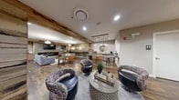 Located at 622 S Broadway in Baltimore, Maryland, this single-floor community lounge offers a well-appointed gathering space that blends rustic charm with modern design. The space is characterized by rich hardwood flooring, exposed reclaimed wood beams, and accent walls featuring a mix of weathered wood planks and textured mosaic tile, creating a warm yet contemporary aesthetic throughout.

The main lounge area is anchored by a sleek fireplace with a mosaic tile surround, paired with a mounted flat-screen television. Comfortable seating options include a grey sofa adorned with colorful accent pillows, a leather lounge chair, and an assortment of stylish swivel chairs arranged around a central coffee table — ideal for casual gatherings or relaxed conversation.

A dedicated kitchenette area provides a concrete-topped bar with bar stool seating, warm wood cabinetry, a stainless steel sink, microwave, dishwasher, and a coffee maker, making the space well-equipped for entertaining. Pendant lighting with globe fixtures illuminates this zone, adding an elegant touch.

Natural light filters through glass-paneled entry doors, complemented by recessed ceiling lighting and pendant fixtures throughout. The space also includes a secondary room currently utilized for package storage. Overall, the lounge delivers a cohesive, inviting atmosphere suited for community use.