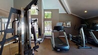 Located at 3952 Boxstead Common in Fremont, California, this fitness center is a thoughtfully designed, single-level space built to support a comprehensive workout experience. The room features vaulted ceilings that rise dramatically toward a central peak, lending a spacious, airy quality enhanced by ceiling fans and recessed lighting throughout.

The space is outfitted with a full suite of cardio equipment, including multiple treadmills, elliptical trainers, a stair climber, and a recumbent bike — accommodating a range of fitness preferences. A multi-station cable machine serves as the centerpiece for strength training, positioned alongside a dedicated bench and free weight area. Full-length mirrors along one wall expand the visual depth of the room and allow users to monitor their form during exercise.

Natural light enters through multiple windows on several walls, offering views of surrounding greenery and an outdoor area that appears to include a patio or pool space. Neutral taupe and dark brown walls complement the carpeted flooring, creating a focused yet inviting environment. Decorative touches such as potted plants and framed athletic artwork add warmth to the functional setting, rounding out a well-balanced fitness facility.