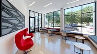Located at 2202 S Kinnickinnick, this single-floor commercial space offers a striking blend of modern architecture and thoughtful interior design. The entrance features black-framed floor-to-ceiling glass doors and walls that flood the lobby with natural light, providing clear views of the surrounding tree-lined streetscape. Polished terrazzo flooring in warm neutral tones extends throughout the space, lending a clean and refined aesthetic underfoot.

The lobby and reception area are appointed with iconic red egg chairs paired with a sleek white side table, set against crisp white walls adorned with a large-scale wave-pattern acoustic art panel. This design motif is carried through multiple areas of the space, reinforcing a cohesive visual identity. Recessed linear LED lighting runs across the ceilings, providing consistent, modern illumination.

A spacious lounge and collaborative area features an expansive black-framed window wall that frames outdoor greenery, filling the interior with abundant natural light. Built-in bench seating along the window line is complemented by modular tables and modern chairs, creating a versatile setting for work or meetings. Glass-partitioned interior rooms are visible from the main area, maintaining an open and transparent feel while defining functional zones. The overall ambiance is clean, bright, and professionally designed.