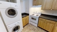 This single-floor space is thoughtfully designed to maximize functionality within a compact layout. The kitchen is outfitted with light natural wood cabinetry that provides ample storage through both upper and lower cabinet configurations. Dark granite countertops offer a striking contrast against the warm wood tones, lending a practical yet polished aesthetic to the cooking area. A white electric coil-top range with an overhead ventilation hood serves as the focal point of the cooking zone, complemented by additional counter space on either side. The flooring throughout features neutral-toned large-format ceramic tiles, contributing to a clean and cohesive look that is easy to maintain. A notable feature of this space is the seamlessly integrated stacked washer and dryer unit, tucked into a dedicated nook adjacent to the kitchen. This in-unit laundry solution adds significant convenience without occupying excessive floor space. The overall design balances practicality with a tidy, understated aesthetic, making efficient use of every available area within the single-floor layout.