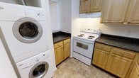 This single-floor space is thoughtfully designed to maximize functionality within a compact layout. The kitchen is outfitted with light natural wood cabinetry that provides ample storage through both upper and lower cabinet configurations. Dark granite countertops offer a striking contrast against the warm wood tones, lending a practical yet polished aesthetic to the cooking area. A white electric coil-top range with an overhead ventilation hood serves as the focal point of the cooking zone, complemented by additional counter space on either side. The flooring throughout features neutral-toned large-format ceramic tiles, contributing to a clean and cohesive look that is easy to maintain. A notable feature of this space is the seamlessly integrated stacked washer and dryer unit, tucked into a dedicated nook adjacent to the kitchen. This in-unit laundry solution adds significant convenience without occupying excessive floor space. The overall design balances practicality with a tidy, understated aesthetic, making efficient use of every available area within the single-floor layout.