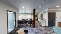 This thoughtfully designed single-floor amenity space blends functionality with contemporary style, offering a variety of settings suited for collaboration, relaxation, and productivity. The open layout is anchored by a central conference area featuring a substantial wood-topped table surrounded by adjustable task chairs, ideal for meetings or group work sessions. A structural wood column adds warmth and visual interest to the space, complementing the rich wood trim that frames the doors and cabinetry throughout.

Adjacent to the conference area, a comfortable lounge zone provides plush seating with a geometric wood coffee table, curated wall art, and access to an exterior-facing window that brings in natural light. A sliding barn-style door with glass panels opens to a bright, windowed lounge overlooking a railing — flooding the area with daylight and offering views of the surrounding urban environment. This sun-drenched nook features additional lounge chairs and natural wood stump accent tables.

The space also includes a dedicated private meeting room, a shared printer station with a white storage cabinet, and a neatly styled credenza nook with a textured brick accent wall. Geometric patterned carpeting flows throughout, tying together the modern aesthetic. Recessed square lighting fixtures illuminate the space cleanly and evenly, completing the polished, professional atmosphere.