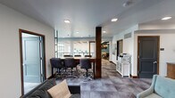 This thoughtfully designed single-floor amenity space blends functionality with contemporary style, offering a variety of settings suited for collaboration, relaxation, and productivity. The open layout is anchored by a central conference area featuring a substantial wood-topped table surrounded by adjustable task chairs, ideal for meetings or group work sessions. A structural wood column adds warmth and visual interest to the space, complementing the rich wood trim that frames the doors and cabinetry throughout.

Adjacent to the conference area, a comfortable lounge zone provides plush seating with a geometric wood coffee table, curated wall art, and access to an exterior-facing window that brings in natural light. A sliding barn-style door with glass panels opens to a bright, windowed lounge overlooking a railing — flooding the area with daylight and offering views of the surrounding urban environment. This sun-drenched nook features additional lounge chairs and natural wood stump accent tables.

The space also includes a dedicated private meeting room, a shared printer station with a white storage cabinet, and a neatly styled credenza nook with a textured brick accent wall. Geometric patterned carpeting flows throughout, tying together the modern aesthetic. Recessed square lighting fixtures illuminate the space cleanly and evenly, completing the polished, professional atmosphere.