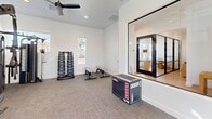 Located at 9975 W Encanto Blvd in Avondale, Arizona, this fitness center delivers a comprehensive workout experience within a thoughtfully designed single-room layout. The space features crisp white walls, neutral carpet flooring, and vaulted ceilings fitted with dual ceiling fans that promote airflow and comfort during exercise sessions. Abundant natural light streams in through multiple large windows adorned with white blinds, offering views of the surrounding outdoor area including what appears to be a pool and palm trees — adding a bright, resort-like atmosphere to every workout. The cardio zone is anchored by multiple treadmills and a stair climber machine, all positioned to take advantage of the generous window views. On the strength training side, the space is equipped with a fully loaded multi-functional rack system featuring medicine balls, kettlebells, battle ropes, resistance bands, weight plates, and a cable machine attachment — providing a wide range of training options. A dumbbell rack with a flat utility bench rounds out the free weights area. A large glass window wall connects the fitness room to an adjacent interior space, adding visual openness. Recessed lighting and a clean, minimalist design make this fitness center both functional and inviting.