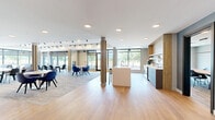 Located at 1994 Springfield Rd in Kelowna, BC, this thoughtfully designed family room is a contemporary shared social space that seamlessly integrates a kitchen, dining area, and living lounge within a single open-plan floor. Light hardwood flooring flows throughout, complemented by a neutral palette of grey and blue tones, wood-panelled accents, and textured wall finishes that lend warmth and visual depth to the interior. The expansive dining area is a central highlight, anchored by a long wooden communal table beneath a striking branched chandelier. Floor-to-ceiling windows along one wall bathe the space in natural light and offer sweeping outdoor views of the surrounding Kelowna landscape, including mature trees and distant mountains. The fully equipped kitchen features sleek, flat-front grey cabinetry, a white tile backsplash, stainless steel appliances including a double wall oven and French-door refrigerator, and a generous island with bar seating. Adjacent to the dining area, a comfortable living lounge is outfitted with modular seating, vibrant yellow ottoman accents, and a large wall-mounted television — ideal for relaxed gatherings. Glass-panel doors define distinct zones while maintaining an open, airy feel throughout the space.