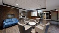 This well-appointed amenity lounge offers a generous, open floor plan spread across a single level, thoughtfully organized into several distinct functional zones. The space is unified by rich, dark hardwood flooring and a drop tile ceiling with recessed and pendant lighting that creates a warm, refined ambiance throughout.

At the entry, a striking accent wall clad in dark wood paneling sets an elegant tone, complemented by wall sconces and a sleek glass console table with decorative greenery. The main lounge area features comfortable upholstered armchairs arranged around a marble-top cocktail table, alongside a bold blue sofa that adds a vibrant accent. A fireplace with a textured brick surround anchors one wall, adding warmth and character.

A dedicated media lounge is outfitted with a large wraparound sectional sofa, round ottomans, and a ceiling-mounted projector, making it ideal for group screenings or casual gatherings. Adjacent to this is a dining area with multiple wood-top tables and upholstered side chairs, illuminated by large drum pendant lights and framed artwork.

The space also includes a pass-through kitchenette with white cabinetry and appliances, along with private nook seating divided by a low booth-style partition. A quieter seating alcove near frosted glass doors provides a more intimate retreat. The overall design balances contemporary style with functional versatility.