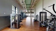 This expansive fitness center occupies a single floor and offers a well-appointed environment for a wide range of workout disciplines. The space is defined by its industrial-chic architectural design, showcasing exposed concrete ceilings with green-painted ductwork, pendant lighting, and structural concrete columns that lend the facility a modern, urban character.

Warm hardwood flooring spans the majority of the exercise area, complemented by rubber flooring in designated strength training zones. Floor-to-ceiling steel-framed windows line multiple walls, flooding the space with natural light and providing open views of the surrounding outdoor environment.

The main exercise floor is generously proportioned and houses a broad selection of professional-grade strength machines, cable systems, power racks, and a dedicated free weights area complete with a full dumbbell rack and adjustable benches. A TRX and kettlebell station is positioned against a window wall, offering a functional training zone with an inspiring outdoor backdrop. Large mirrors enhance the sense of openness in the free weights section.

A separate, more intimate training room provides a quieter setting with its own equipment and wall-mounted display screen. Additional amenities include a built-in storage and equipment counter with shelving, a water station, and wall-mounted screens throughout. The overall ambiance is clean, professional, and energizing.