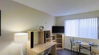 This single-floor space offers a practical and efficiently arranged living area designed to maximize functionality. The room features light-colored walls with a textured ceiling, contributing to a clean and neutral aesthetic. Dark hardwood-style flooring grounds the space and provides a subtle contrast to the light tones throughout.

A built-in light wood workstation serves as a focal point, incorporating an L-shaped desk surface alongside open shelving units with multiple compartments, ideal for storage and display. The shelving accommodates media, books, and decorative items, while a flat-screen television is positioned atop the unit for convenient viewing.

A window dressed with vertical blinds allows natural light to filter into the space, brightening the room considerably. A small dining or accent area is visible near the window, furnished with a metal-framed chair and compact table, offering an additional functional zone within the room.

The overall ambiance is understated and utilitarian, with a layout that prioritizes practicality. The combination of built-in furniture and open shelving makes efficient use of available space, while the neutral palette keeps the environment feeling open and uncluttered.