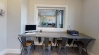 This single-floor business center is thoughtfully designed to support a range of work and collaboration needs in a clean, contemporary setting. The space features neutral gray walls, white trim, and patterned commercial carpeting throughout, creating a professional yet approachable atmosphere.

A wall-mounted counter workstation provides multiple individual work points, each equipped with convenient power outlets and accommodated by industrial-style metal stools. One section of this counter includes a desktop computer and printer, making it a fully functional shared workstation. A pass-through interior window at this station offers a direct sightline into an adjacent glass-walled conference room, which is furnished with a long table and ergonomic chairs — well-suited for meetings and group discussions.

A standing-height bistro table with metal stools is positioned near two large windows, allowing natural light to fill the area and offering views of the surrounding neighborhood. This spot serves as an informal gathering or break area within the space.

Additional storage or utility access is available through a separate door visible from the main area. Tasteful wall art, including cityscape paintings and abstract canvases, adds visual interest and personality to the environment. Overall, the space is efficiently laid out and well-equipped to serve as a productive shared workspace or community business center.