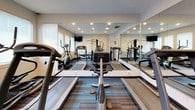 This single-level fitness center is designed to support a well-rounded workout routine, offering a thoughtfully arranged selection of cardio and strength training equipment. The space is outfitted with multiple treadmills, elliptical machines, a stationary bike, and a multi-function cable machine, providing users with a comprehensive range of exercise options.

A full wall of floor-to-ceiling mirrors along one side visually expands the room and allows users to monitor their form during workouts. Recessed lighting throughout the ceiling provides consistent, even illumination, while a row of windows fitted with horizontal blinds along the perimeter wall introduces natural light and adds an open, airy quality to the space.

The flooring features a contemporary patterned carpet with teal and gray tones, adding a modern aesthetic to the functional environment. Mounted flat-screen televisions are positioned for visibility from the cardio equipment, enhancing the workout experience. A glass-paned interior door connects the fitness area to an adjacent tiled space, and a sanitizing station with waste disposal is conveniently located near the entrance, promoting cleanliness and hygiene throughout.