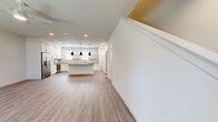 Located in Buda, TX, this two-story home offers a clean, modern aesthetic with thoughtful design across both floors. The main level hosts an open-concept kitchen and living area, filled with natural light from large windows that frame neighborhood views. The kitchen is a standout feature, showcasing white shaker cabinetry, open floating shelves, a subway tile backsplash, a curved center island with dark countertops, stainless steel appliances, and bold black pendant lighting — all complemented by warm wood-look flooring that flows throughout the home. The living area is spacious and inviting, with a ceiling fan and a front door entry with transom window adding brightness. A staircase leads to the upper level, where two bedrooms offer comfortable retreats, each with natural light from well-positioned windows. The home features four bathrooms in total, including one with a walk-in glass-enclosed shower finished with large-format tile, and another with a full soaking tub and tiled surround. All bathrooms feature wood-look flooring, white vanities with dark hardware, and crisp, neutral finishes. Outdoors, a shared green space with manicured turf, landscaping, and a covered pergola area adds to the community's appeal.