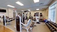 Located at 659 Rice Boulevard in Exton, Pennsylvania, this fitness center offers a well-appointed, single-floor workout facility designed to support a comprehensive exercise routine. The space features a clean, neutral interior with a drop-panel ceiling equipped with recessed fluorescent lighting that ensures bright, even illumination throughout. Large wall mirrors enhance the sense of openness and allow users to monitor their form during workouts. The fitness area is outfitted with a solid selection of strength training equipment, including a prominent multi-station cable and weight machine, a dedicated weight bench, and a variety of free weights. On the cardio side, the facility provides multiple treadmills, an upright stationary bike, and additional aerobic machines, all positioned to allow comfortable spacing between users. Stability balls and supplemental fitness accessories round out the available equipment. The flooring is low-pile carpet suited for fitness use, and the room features neutral-toned walls that contribute to a focused, no-distraction workout environment. Emergency exit access is clearly marked, and the space is neatly organized, reflecting a well-maintained facility. Overall, this fitness center provides a functional and inviting environment for residents or members seeking a convenient on-site exercise option.