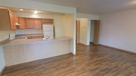 This single-floor space showcases an open-concept layout that seamlessly connects the kitchen and living area, creating a functional and cohesive environment. The kitchen is equipped with warm honey-toned wood cabinetry, a white refrigerator, and a built-in range. A peninsula with a light wood-finish countertop and an integrated sink extends into the living space, providing both additional workspace and a casual dining or seating option as a breakfast bar.

The living area is finished with rich, dark hardwood-style flooring that extends throughout, adding warmth and visual continuity to the space. Light-colored walls contribute to a bright, open atmosphere. A structural column near the peninsula defines the boundary between the kitchen and living zones while preserving the open feel of the layout.

Visible toward the rear of the space are doorways leading to additional rooms, including what appears to be a closet with bi-fold doors, offering practical storage solutions. A wall-mounted thermostat is also visible, indicating individual climate control. The overall design is practical and straightforward, with a neutral palette suited to a variety of interior styles.