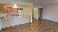 This single-floor space showcases an open-concept layout that seamlessly connects the kitchen and living area, creating a functional and cohesive environment. The kitchen is equipped with warm honey-toned wood cabinetry, a white refrigerator, and a built-in range. A peninsula with a light wood-finish countertop and an integrated sink extends into the living space, providing both additional workspace and a casual dining or seating option as a breakfast bar.

The living area is finished with rich, dark hardwood-style flooring that extends throughout, adding warmth and visual continuity to the space. Light-colored walls contribute to a bright, open atmosphere. A structural column near the peninsula defines the boundary between the kitchen and living zones while preserving the open feel of the layout.

Visible toward the rear of the space are doorways leading to additional rooms, including what appears to be a closet with bi-fold doors, offering practical storage solutions. A wall-mounted thermostat is also visible, indicating individual climate control. The overall design is practical and straightforward, with a neutral palette suited to a variety of interior styles.