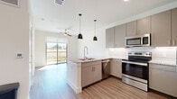 This well-designed 1-bedroom, 1-bathroom residence in Austin, TX offers a thoughtful open-concept layout that maximizes every inch of its footprint. The combined kitchen and living area serves as the heart of the home, anchored by a peninsula with granite countertops and a built-in sink, illuminated by a pair of stylish pendant lights. The kitchen is equipped with stainless steel appliances—including a range, microwave, and dishwasher—complemented by sleek taupe cabinetry with under-cabinet lighting and a classic subway tile backsplash. Wood-look flooring runs throughout, adding warmth and continuity. The living area benefits from abundant natural light and direct access to a private balcony through sliding glass doors, offering outdoor views of the surrounding greenery. A ceiling fan overhead enhances comfort in the main living space. The bedroom features dual windows that invite natural light and views of the outdoors. Three closets provide generous storage throughout the unit. The bathroom is finished with a granite-topped vanity, a large mirror, wood-look tile flooring, and a glass-enclosed shower. Clean white walls and modern finishes create a bright, contemporary ambiance throughout.