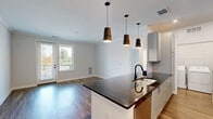 Located at 4541 Helm Dr in Virginia Beach, Virginia, this single-floor residence offers a well-designed, open-concept layout with modern finishes throughout. The kitchen serves as a central focal point, boasting dark granite countertops on a spacious island with an undermount sink and matte black fixtures. Gray shaker-style cabinetry lines the walls, complemented by stainless steel appliances including a refrigerator, range, and built-in microwave. Pendant lighting in a bold, dark finish hangs above the island, adding a contemporary touch. The kitchen flows seamlessly into the living area, which features hardwood-style flooring, crown molding, and a glass door leading to a private balcony with outdoor views. The bedrooms are carpeted and filled with natural light from large windows with views of the surrounding landscape. A dedicated laundry area houses a washer and water heater. The bathroom is clean and modern, with white shaker vanity cabinetry, a quartz countertop, a large mirror, matte black hardware, and a tub-shower combination. Additional rooms with hardwood-style flooring and crown molding offer flexible living space. Neutral tones and consistent finishes create a cohesive, move-in-ready ambiance throughout the home.