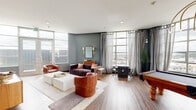 This sophisticated amenity lounge is designed to impress, offering a well-appointed, open-plan space that seamlessly blends recreation, relaxation, and social gathering. Large floor-to-ceiling windows wrap around the room, flooding the interior with natural light and framing sweeping panoramic city views that serve as a striking backdrop throughout.

The recreational zone anchors the space with a classic billiard table crafted from rich hardwood, positioned beneath a sculptural gold arched chandelier that adds a refined, artistic focal point. Sheer gray curtains frame the expansive windows, softening the natural light while maintaining the airy atmosphere.

The lounge area features warm, earthy tones with a channeled plum velvet sofa, cognac leather armchairs, and a clean white coffee table set against a muted slate-blue accent wall. Brass wall sconces and a vintage-style wooden credenza with a mirror add warmth and depth to this intimate seating vignette.

A dining and gaming zone offers a sleek dark table surrounded by black-and-gold chairs, with an adjacent wall-mounted screen. The kitchenette area is a design standout, featuring vibrant green cabinetry with brass hardware, a bold geometric black-and-white tiled backsplash, and counter space equipped with appliances. All-gender restrooms complete the space, finished with matching green cabinetry and the same eye-catching patterned tile. Warm wood-look flooring ties every zone together cohesively.