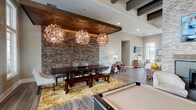Located at 410 Starmount Park Blvd in Kannapolis, NC, this single-floor clubhouse is a thoughtfully designed community gathering space that blends upscale finishes with welcoming functionality. The main lounge area makes a strong architectural statement with soaring stacked-stone accent walls, exposed dark wood ceiling beams, and wood-plank tile flooring that runs throughout the space. A floor-to-ceiling stone fireplace anchors one end of the room, flanked by white lounge chairs and a wall-mounted flat-screen TV, creating an inviting focal point. A regulation billiards table occupies the center of the space beneath an ornate globe chandelier, while a large dining table with bench seating is set beneath a trio of sculptural pendant lights backed by a striking stone feature wall with warm accent lighting. Multiple seating clusters are positioned throughout the open-plan layout to encourage conversation and relaxation. The full kitchen is outfitted with dark cabinetry, granite countertops, stainless steel appliances, pendant lighting, and a breakfast bar with upholstered swivel seating. Oversized windows and glass French doors along one wall flood the interior with natural light while offering direct views of the outdoor pool and patio area, seamlessly connecting the indoor and outdoor amenities.