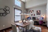 This thoughtfully designed single-floor space seamlessly blends a sophisticated dining area with a functional built-in workstation, creating a versatile and visually compelling interior. The soaring ceilings lend an airy, expansive quality to the main area, while large windows invite generous natural light and frame outdoor views of lush greenery beyond dark-framed French doors.

The dining area serves as the centerpiece of the space, anchored by a striking reclaimed wood table surrounded by white upholstered chairs with chrome accents. A bold industrial-style pendant light fixture hangs overhead, adding character and warmth. A patterned area rug in earthy tones grounds the seating arrangement, complementing the rich hardwood flooring that flows throughout.

Integrated seamlessly along one wall is a built-in workstation featuring an extended granite-topped countertop with dark cabinetry below. The workspace is enhanced by wall-mounted sconce lighting and a series of recessed niche shelves displaying decorative ceramic vessels, adding an artistic touch. Abstract artwork panels adorn the adjacent wall, further elevating the design aesthetic.

Neutral grey walls provide a cohesive backdrop that ties together the warm wood tones, chrome fixtures, and curated décor. The overall ambiance is modern yet welcoming, with a refined balance of practicality and style.