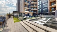 Located at 1395 E Miller Ave in Millcreek, Utah, this expertly designed outdoor amenity space is set against the backdrop of a modern multi-story residential building clad in brick, warm wood tones, and sleek metal paneling. The courtyard level centers around a resort-style shallow pool with clean concrete coping, stainless steel handrails, and an adjacent hot tub spa, all framed by lounge chairs and large cantilever umbrellas for shade. Lush turf areas are interspersed with paved walkways, providing comfortable seating zones with stylish wicker furniture arranged for both relaxation and socializing. A covered pergola structure offers a shaded gathering spot alongside the greenery. Raised planter beds featuring ornamental grasses and columnar evergreen trees line seating areas finished in polished concrete and decorative stone gravel, adding a refined, landscaped aesthetic to the hardscape. A rooftop terrace level crowns the amenity experience, offering sweeping panoramic views of the Wasatch Mountains to the east, furnished with lounge chairs, seating groups, and artificial turf sections ideal for outdoor gatherings. The entire space benefits from abundant natural light and open sky exposure, creating a vibrant, resort-caliber outdoor environment in the heart of Millcreek.