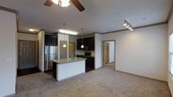 This single-floor residence showcases an open-concept layout that connects the kitchen and living area into one cohesive, functional space. The kitchen is a standout feature, equipped with dark espresso cabinetry that extends to the upper walls, providing ample storage while delivering a bold, contemporary aesthetic. A central island with granite countertops and an integrated sink serves as both a prep area and a casual gathering point, illuminated by pendant lighting that hangs from the ceiling above. Stainless steel appliances, including a full-size refrigerator and built-in microwave, complement the dark cabinetry. Track lighting near the adjoining area adds additional illumination, while a ceiling fan overhead enhances comfort. The living area transitions seamlessly from the kitchen, featuring neutral carpet flooring that contrasts with the dark kitchen tile, creating a defined yet open feel. The light-toned walls and crown molding add a refined touch throughout. A doorway visible from the main space leads to additional rooms, suggesting further private quarters. Overall, the space presents a clean, modern design with practical amenities and a well-balanced aesthetic.
