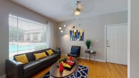 This single-story residence offers a well-organized layout that balances comfort and functionality throughout its main living areas. The living room serves as the heart of the space, featuring warm hardwood-style flooring, neutral gray walls, and a large window dressed with horizontal blinds and sheer curtains that frame a view of an outdoor pool area. A ceiling fan with light fixture provides overhead comfort, while a bold blue-and-yellow abstract canvas and a patterned area rug add a vibrant, contemporary touch to the room.

The galley-style kitchen is efficiently designed with light wood cabinetry, dark granite-style countertops, and black appliances including a range, microwave, refrigerator, and dishwasher. The same hardwood-style flooring continues throughout, and a door at the end of the kitchen leads to a dedicated laundry area.

The bedroom is carpeted for comfort and includes a mirrored sliding-door closet that enhances the sense of space. A window with blinds allows natural light to filter in, and the room provides ample space for both a sleeping area and a desk workspace.

The bathroom features hardwood-style flooring, a vanity with dark countertop and wood cabinetry, and a full tub-and-shower combination accented with classic black tile detailing. The overall ambiance of the residence is modern, practical, and inviting.