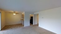 This single-floor residence offers a practical and comfortable layout designed for everyday living. The main living area is carpeted in neutral tones, creating a warm and inviting atmosphere. A ceiling fan with an integrated light fixture adds both function and a casual aesthetic to the space.

The living area flows openly toward the kitchen, which is partially separated by a breakfast bar — ideal for casual dining or entertaining. The kitchen features light-toned cabinetry, tile flooring, and black appliances, including a refrigerator and range, providing a clean contrast to the lighter surroundings. A hallway corridor connects the living area to additional rooms beyond, contributing to the overall sense of an organized and navigable floor plan.

The walls throughout are finished in a clean, neutral white, maximizing the sense of light and openness. A thermostat is visibly mounted on the living room wall, indicating climate control throughout the unit. Tile flooring in the kitchen transitions seamlessly to carpet in the living space, clearly defining each zone. Overall, the space presents a straightforward, functional design with a simple, clean aesthetic well-suited for comfortable day-to-day living.