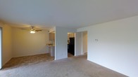 This single-floor residence offers a practical and comfortable layout designed for everyday living. The main living area is carpeted in neutral tones, creating a warm and inviting atmosphere. A ceiling fan with an integrated light fixture adds both function and a casual aesthetic to the space.

The living area flows openly toward the kitchen, which is partially separated by a breakfast bar — ideal for casual dining or entertaining. The kitchen features light-toned cabinetry, tile flooring, and black appliances, including a refrigerator and range, providing a clean contrast to the lighter surroundings. A hallway corridor connects the living area to additional rooms beyond, contributing to the overall sense of an organized and navigable floor plan.

The walls throughout are finished in a clean, neutral white, maximizing the sense of light and openness. A thermostat is visibly mounted on the living room wall, indicating climate control throughout the unit. Tile flooring in the kitchen transitions seamlessly to carpet in the living space, clearly defining each zone. Overall, the space presents a straightforward, functional design with a simple, clean aesthetic well-suited for comfortable day-to-day living.