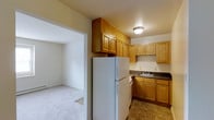 This single-floor residential unit offers a straightforward and functional layout designed for comfortable everyday living. The kitchen is well-equipped with an abundance of warm-toned wooden cabinetry, including upper and lower cabinet storage that provides generous space for organization. A full-size refrigerator, dishwasher, and a double-basin sink are seamlessly integrated into the layout, complemented by dark laminate countertops. The tile flooring in the kitchen adds durability and ease of maintenance. An open pass-through connects the kitchen directly to the adjoining living area, which features soft wall-to-wall carpeting and neutral white walls that create a clean, versatile backdrop. The living space is bathed in ambient ceiling light and offers a welcoming atmosphere. A carpeted bedroom is accessible through an open doorway, revealing a sizable closet with classic bifold doors, offering practical storage solutions. Flush-mount ceiling fixtures provide consistent lighting throughout the unit. The neutral color palette and efficient use of space make this unit a practical and move-in ready home.