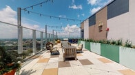 Located atop a high-rise at 850 S Fourth St in Louisville, Kentucky, this rooftop terrace offers a well-appointed outdoor experience with sweeping panoramic views of the city skyline and surrounding neighborhoods. The space is defined by its distinctive checkerboard-patterned tile flooring in warm earth tones and terrazzo-style finishes, lending a stylish, contemporary character to the entire terrace.

The open-air section features multiple seating arrangements centered around a mosaic-tiled fire table, surrounded by wooden lounge chairs with cushioned seating. Festoon string lights are strung overhead, creating an inviting ambiance for evening gatherings. Glass panel railings line the perimeter, ensuring unobstructed views of the Louisville cityscape while maintaining a sense of openness and safety at elevation.

A covered lounge area, sheltered beneath a decorative laser-cut metal pergola with intricate botanical patterns, provides a more intimate setting. This zone includes wingback chairs with turquoise accent cushions, a wall-mounted television, and a built-in fireplace, making it suitable for year-round use. A built-in bench with bold blue-and-white striped cushions runs along the glass railing, ideal for taking in the expansive city views.

Verdant planters in mint green line one wall, filled with ornamental grasses and flowering plants, adding color and a touch of greenery to the urban setting. Audio speakers and lighting fixtures are integrated throughout, completing this thoughtfully designed rooftop retreat.