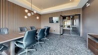 Located at 2274 Princess St in Kingston, Ontario, this professionally designed boardroom suite offers a refined and functional space ideal for meetings and collaborative work. The suite comprises a stylish hallway entrance and a sophisticated conference room, all finished to a high standard.

The hallway sets a polished tone upon entry, featuring a patterned geometric carpet, dark-framed glass doors, and deep grey walls accented by framed black-and-white artwork. The transitions between spaces are seamless, with glass-paneled doors allowing visibility and a sense of openness throughout.

The conference room is the centerpiece of the suite, designed with clear attention to detail. Vertical wood slat wall panels lend warmth and texture, complementing the grey tones of the carpet and walls. A tray ceiling with recessed lighting and a sculptural globe pendant chandelier adds an elevated ambiance to the room. A large wall-mounted display screen serves as a focal point for presentations, while a long boardroom table surrounded by executive leather chairs provides ample seating for meetings. Expansive floor-to-ceiling windows flood the room with natural light and offer views of the surrounding area, creating an bright and energizing atmosphere. A credenza along the wall adds practical storage, completing this well-equipped, contemporary professional space.