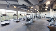 Located at 1900 Southwest 8th Street in Miami's vibrant Little Havana neighborhood, this well-equipped fitness center offers a generous, single-floor layout designed to accommodate a full range of workout needs. The space features an industrial-modern aesthetic, with exposed ductwork and structural elements painted in dark tones against a high ceiling, complemented by pendant lighting that adds a refined touch to the utilitarian design. Patterned commercial carpet in blue and grey tones runs throughout, providing both durability and visual interest underfoot. Floor-to-ceiling windows line the perimeter, flooding the space with abundant natural light and offering sweeping views of the surrounding tree canopy — creating an energizing, open atmosphere. The main workout area is outfitted with a comprehensive selection of Cybex Prestige cardio machines, including treadmills, ellipticals, and stair climbers, all positioned to take full advantage of the outdoor views. A dedicated strength training zone houses a robust array of Cybex selectorized machines, free weights, a dumbbell rack, and functional training equipment. A separate, semi-enclosed area provides space for stretching, core work, and small-group training, complete with stability balls, benches, and wall-mounted TVs. Full-length mirrors enhance spatial perception throughout the strength training zones.