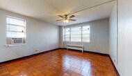 This single-floor, 1-bedroom, 1-bathroom unit offers a straightforward and functional layout designed for comfortable everyday living. The living area is a standout space, featuring warm, richly toned parquet-style tile flooring that adds character throughout. A ceiling fan with an integrated light fixture anchors the room, while a large multi-pane window invites natural light and provides outdoor views. A radiator beneath the window provides reliable heating. The bedroom continues the parquet flooring aesthetic and includes a closet rod for storage, with natural light filtering in from an adjacent window. The kitchen is efficiently arranged in an L-shaped configuration, outfitted with white cabinetry, dark countertops, a double sink, a range, and a full-size refrigerator. Wood-look flooring carries through this space as well. The bathroom features classic white subway-style wall tiles, a tub-shower combination, a pedestal-style vanity with a dark wood cabinet, a mirrored medicine cabinet, and a bold black-and-white checkered floor — adding a timeless, graphic touch. An entryway area with light tile flooring and multiple doors provides organized access to the unit's various rooms and closets.