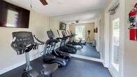 This single-floor fitness center offers a focused and functional workout environment outfitted with a comprehensive selection of modern exercise equipment. The room features multiple elliptical machines and treadmills from True Fitness, providing a range of cardio options for users at any fitness level. A full-wall mirror spans one side of the space, visually expanding the room and allowing users to monitor their form during workouts. The dark rubberized flooring provides a durable, slip-resistant surface well-suited for physical activity. Natural light fills the space through a large window that overlooks an outdoor pool area, creating an inviting and motivating atmosphere. A wall-mounted flat-screen television is positioned for easy viewing during exercise, while a ceiling fan ensures comfortable air circulation throughout. The walls are finished in a warm neutral tone, and white baseboards provide clean, crisp contrast. A fire extinguisher and informational signage near the entry door reflect attention to safety. The overall ambiance is practical yet pleasant, balancing the utility of a well-stocked fitness facility with the comfort of natural light and thoughtful design details.