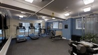 This fitness center offers a practical and well-organized workout environment designed to support a range of exercise routines. The space is finished with blue-painted walls and grey carpet flooring, creating a clean and energizing atmosphere. Overhead fluorescent lighting ensures the room is well-lit throughout, complemented by natural light streaming in through multiple windows that overlook a lush outdoor area.

The cardio section is a highlight, featuring multiple treadmills and an elliptical trainer positioned along the windowed wall, allowing users to enjoy outdoor views during their workouts. A stationary bike and a dedicated weight bench provide additional options for strength and conditioning training. A rack of medicine balls and a set of free weights, including a visible dumbbell rack, round out the strength training offerings.

Exposed piping along the ceiling adds an industrial character to the space, while a traditional radiator along one wall speaks to the building's classic architectural roots. Informational posters and signage are displayed on the walls, contributing to the functional and purposeful design of the room. The overall layout is thoughtfully arranged to maximize usability, making this fitness center a convenient and comprehensive amenity.