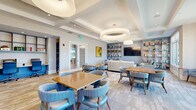 Located at 1231 Arya Way in Ocoee, Florida, this thoughtfully designed library offers a welcoming and versatile communal space across a single floor. The room is characterized by elegant coffered ceilings with large circular and dome pendant light fixtures that provide warm, layered illumination throughout. A soft, neutral color palette of whites, grays, and warm beiges is accented by coordinating blue upholstered seating, creating a cohesive and calming aesthetic.

The space is organized into distinct functional zones. A lounge area features plush sofas and armchairs arranged around a circular side table, anchored by a large flat-screen TV mounted against a wall of built-in shelving filled with books. Adjacent seating vignettes with upholstered accent chairs offer quieter reading nooks. Multiple dining-height tables with curved blue chairs provide space for group activities or individual study.

A standout feature is the sleek wall-mounted linear electric fireplace, which adds a modern touch to a separate seating area. Expansive windows flood the interior with abundant natural light and offer views of the surrounding Florida landscape. Tropical-themed framed artwork adorns the walls, reinforcing the sunny, resort-inspired atmosphere. The combination of hardwood-style and carpeted flooring further defines each zone within this versatile, elegantly finished library.